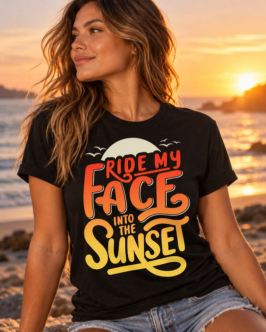 Woman in a black Into the Sunset graphic tee with red-to-yellow sunset lettering, sitting on the beach at sunset