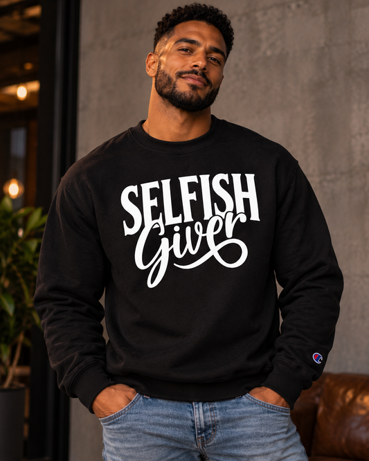 Man in a black Champion crewneck sweatshirt with the white Selfish Giver graphic, hands in his jeans pockets, against a concrete wall